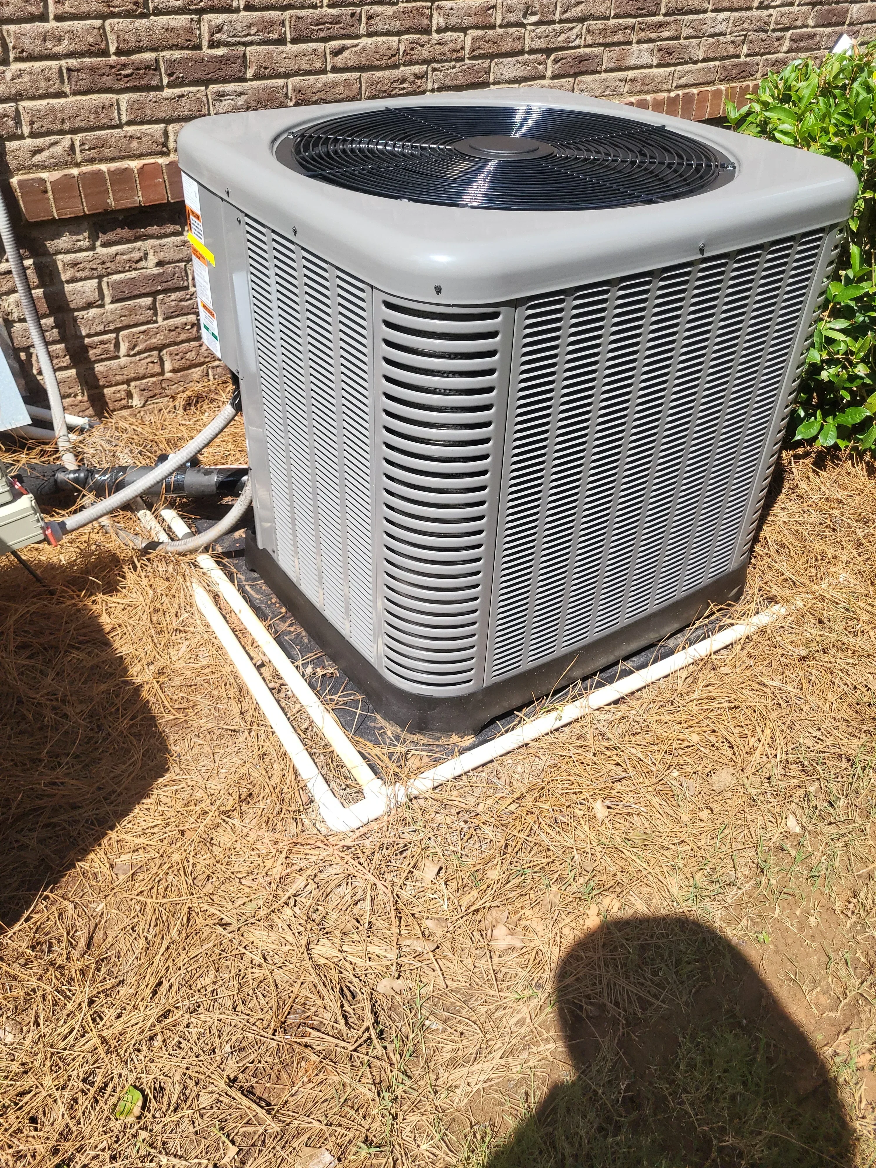 White heat pump condenser unit with black fan and finned coils, mounted on brick foundation with copper refrigerant lines