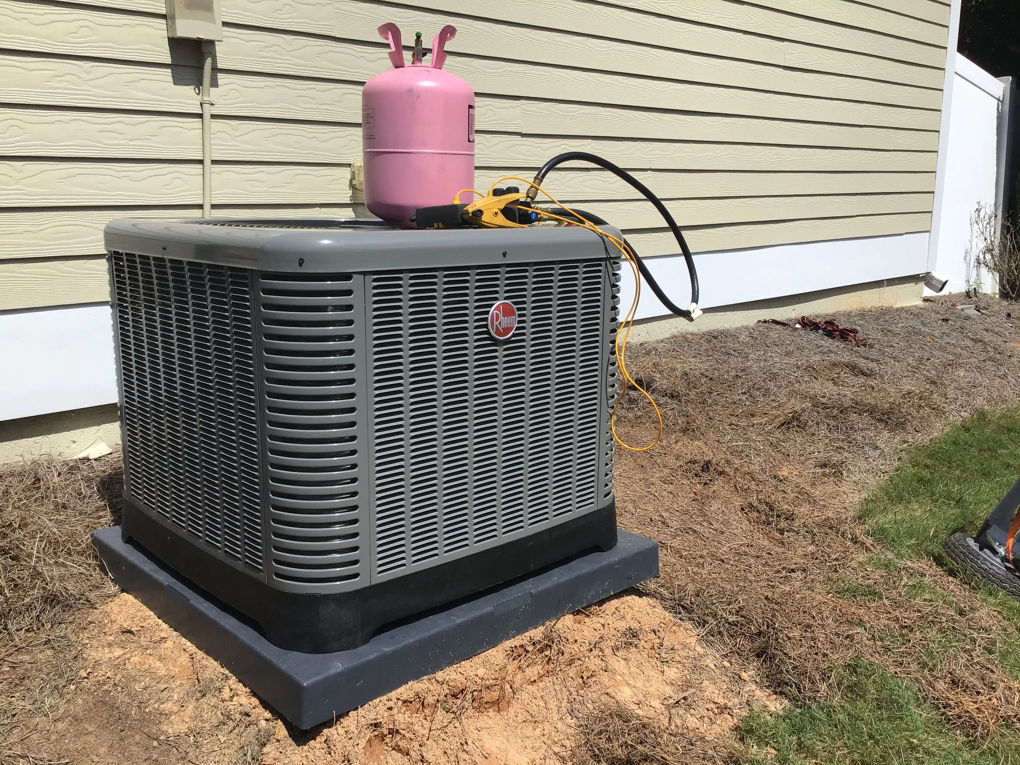 Rheem AC condenser unit on concrete pad with refrigerant cylinder and service equipment