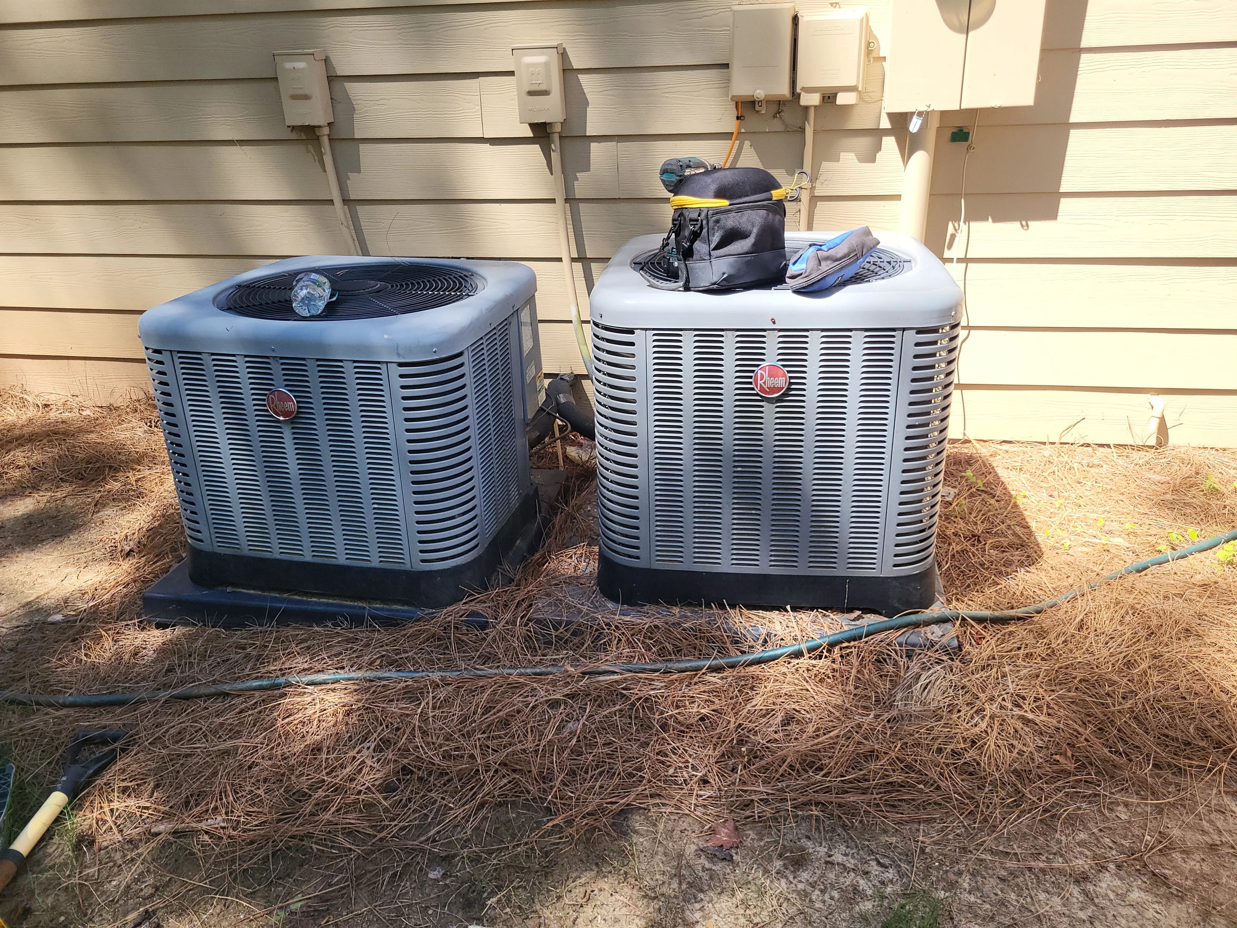 Two Rheem AC condenser units installed at ground level against vinyl siding with electrical connections